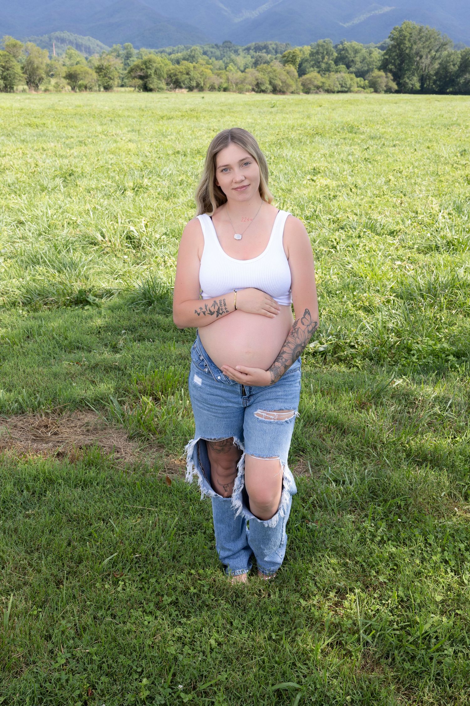Maternity photoshoot in grassy field with mountain backdrop and ripped jeans.