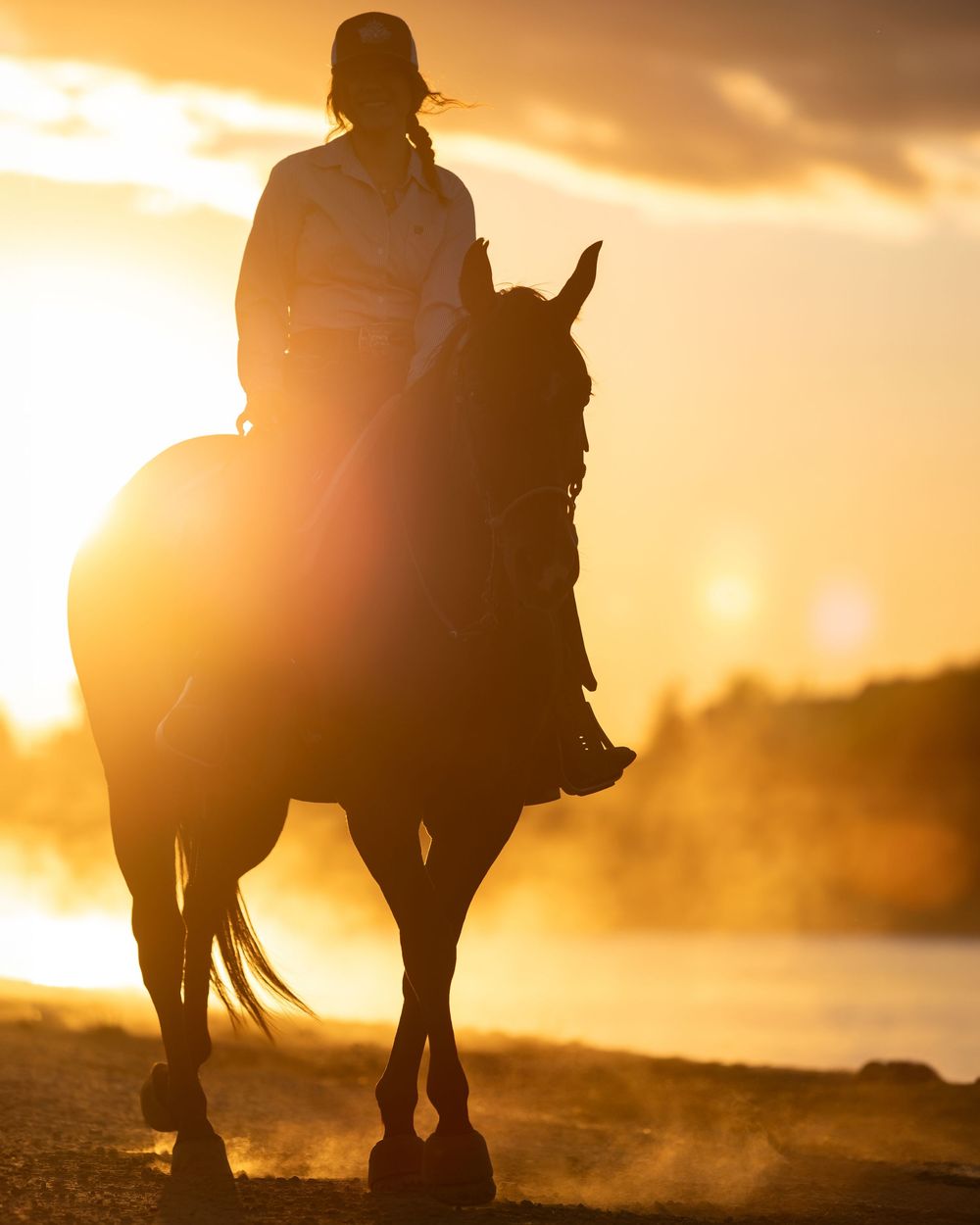 Silhouette of a horse and rider against a stunning golden sunset near a dusty waterfront.