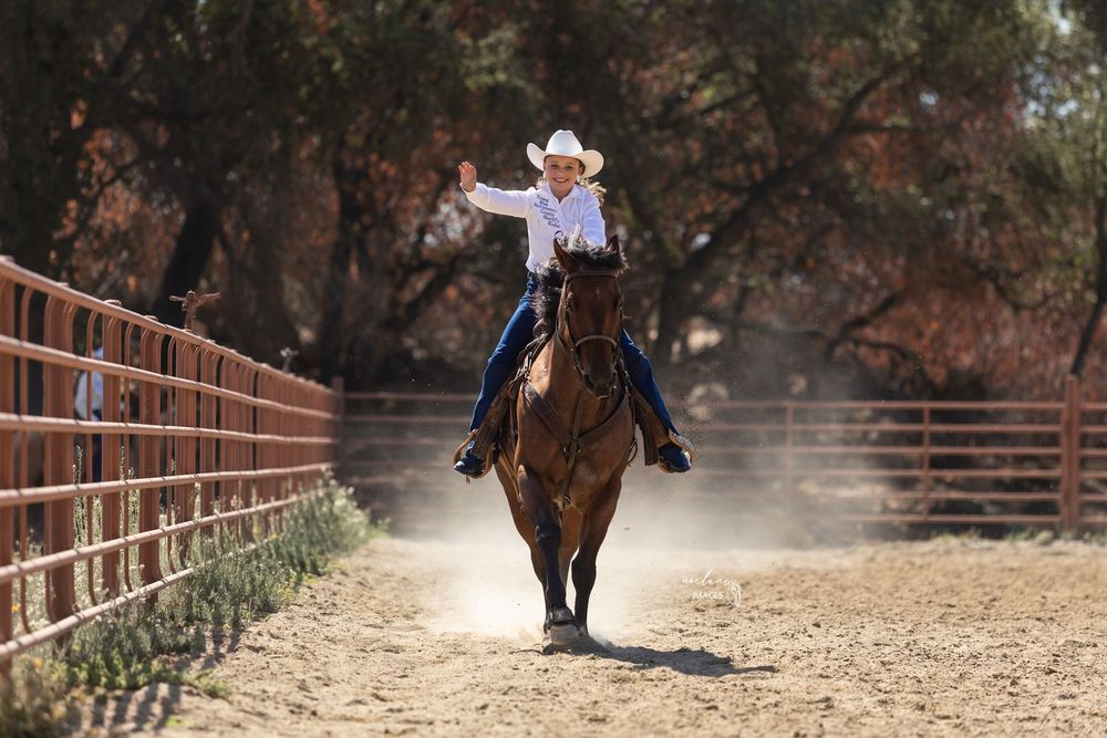 Smiling cowgirl in white hat waving while riding brown horse in fenced ranch arena. Young Miss-Mac Girdlestone-Sac County Sheriffs Rodeo