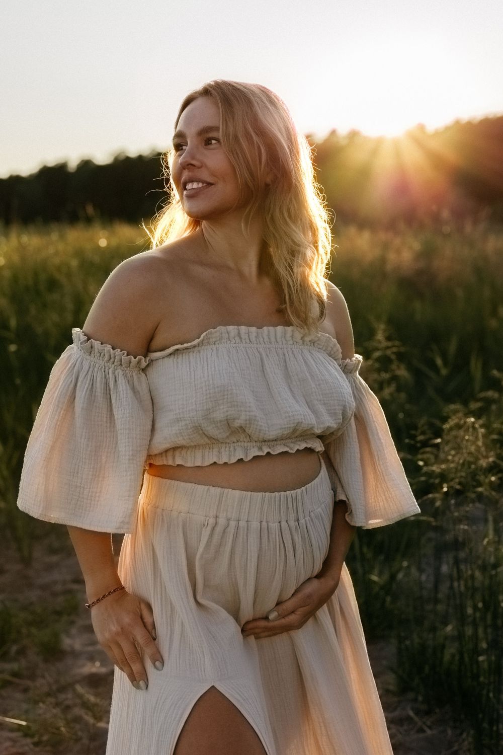 Woman in white off-shoulder dress poses in golden sunset light in summer field.