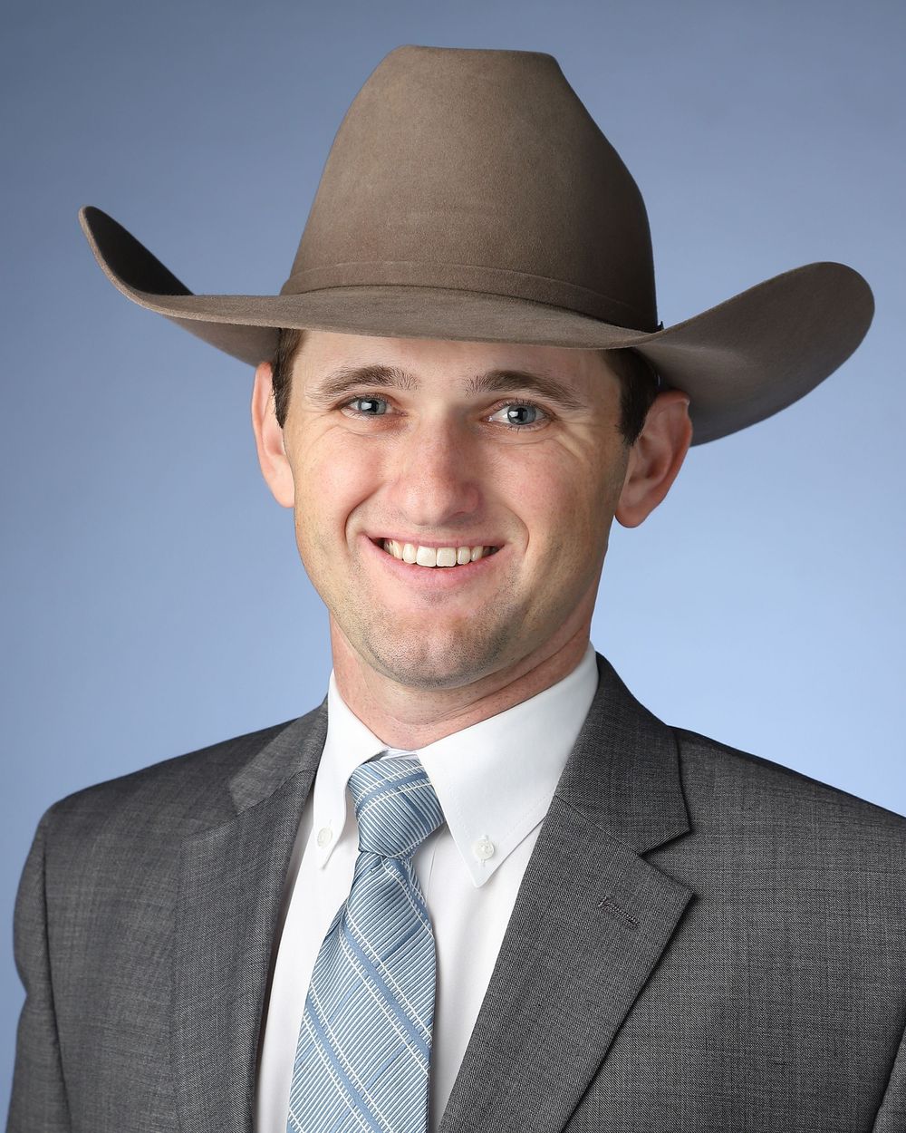 Professional headshot of someone in a gray suit, light blue tie and brown cowboy hat against blue background.