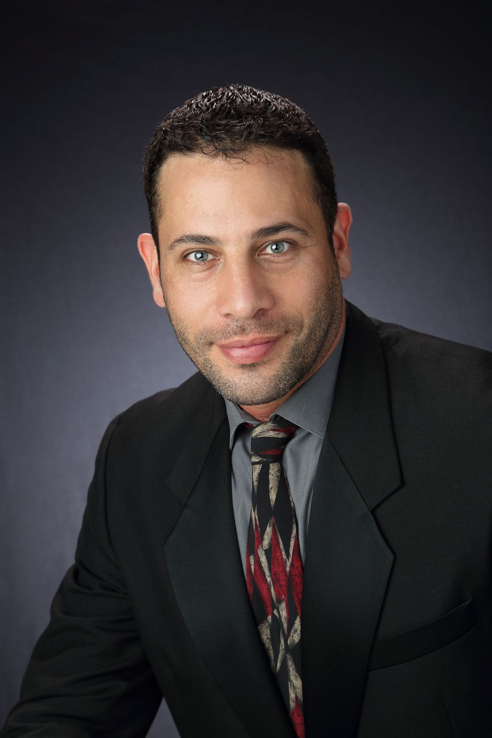 Professional headshot portrait of person in dark suit and patterned tie against gray studio background.
