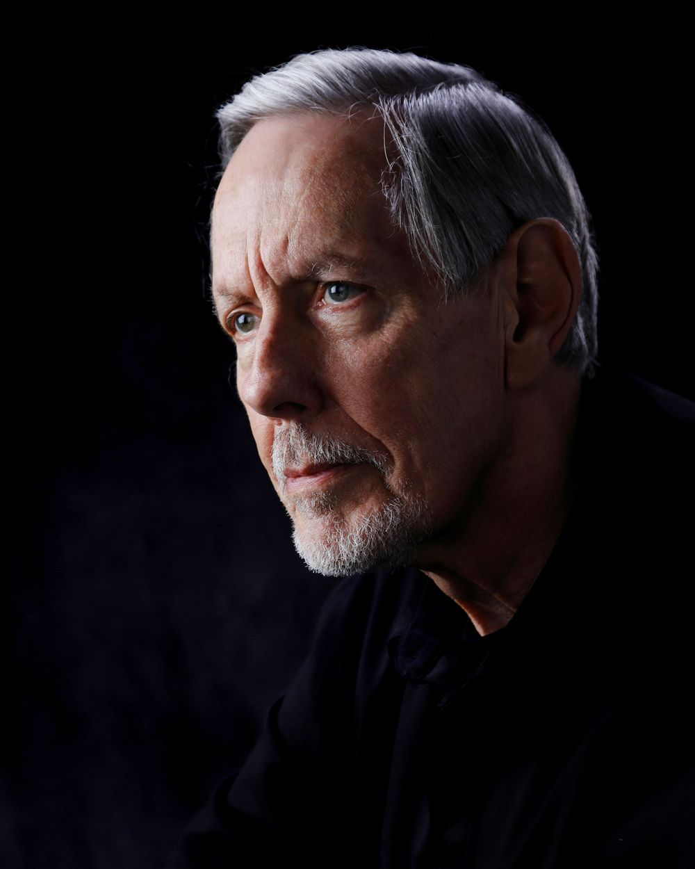 Dramatic side profile portrait in dark lighting with strong shadows against black background.