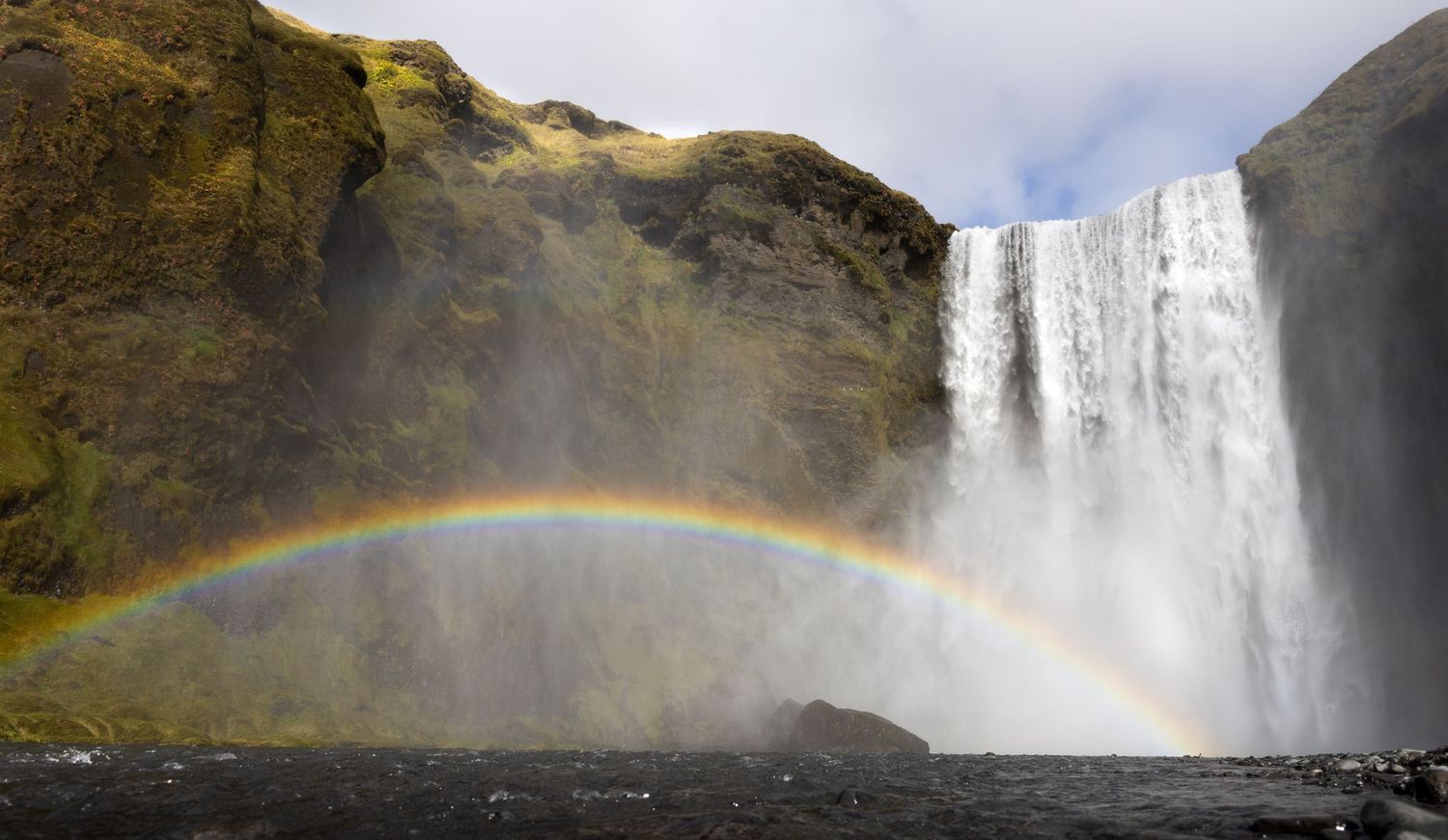 A vibrant rainbow arcs across the misty spray of Skogafoss waterfall in Iceland against dark cliffs and cloudy sky.