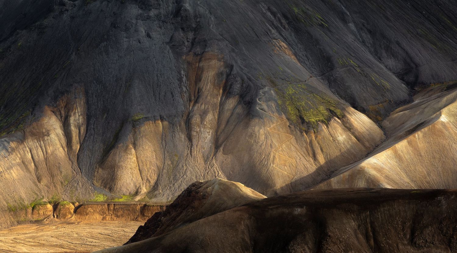 Dramatic sunlight illuminates colorful rhyolite mountains in Landmannalaugar, Iceland against a dark cloudy backdrop.