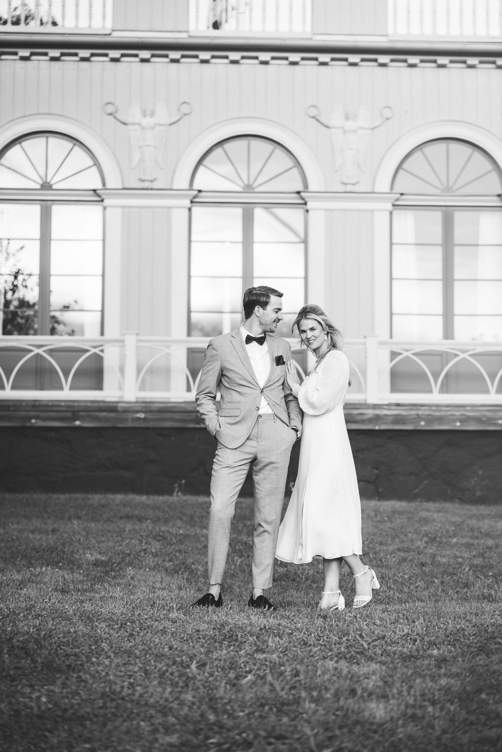 Romantic couple poses on lawn in front of classic white building with arched windows.