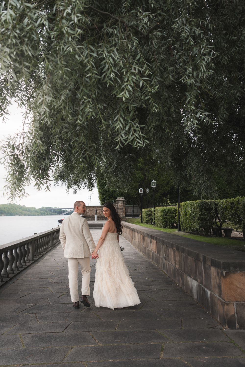 Bride and groom walk hand-in-hand along a waterfront promenade under weeping willow trees in wedding attire.