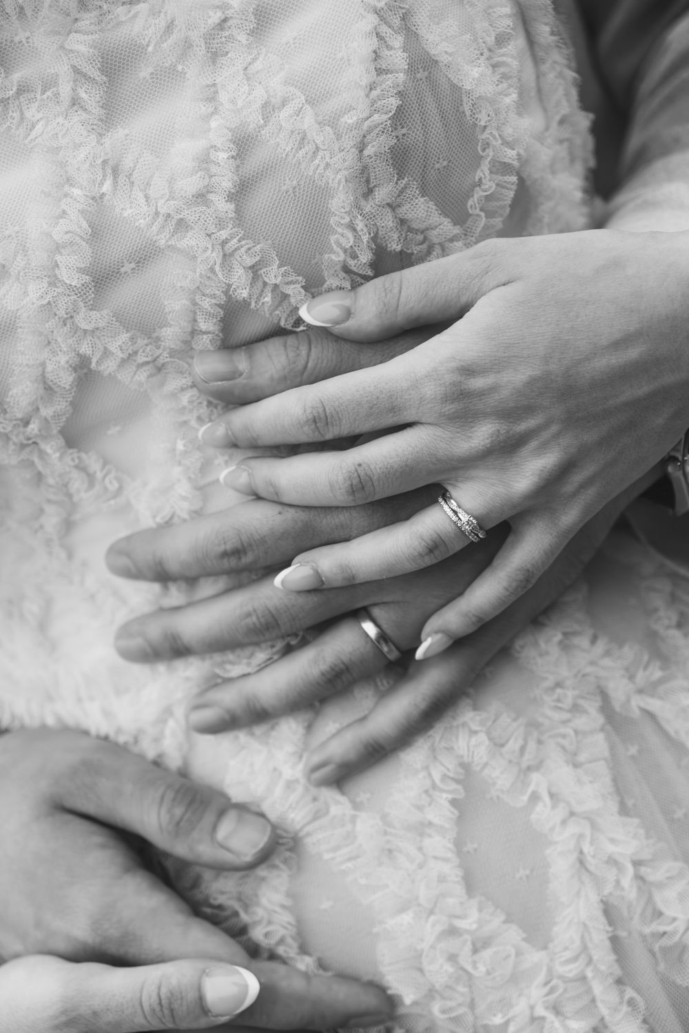 Black and white photo of newlyweds hands with wedding rings resting on brides lace wedding dress.