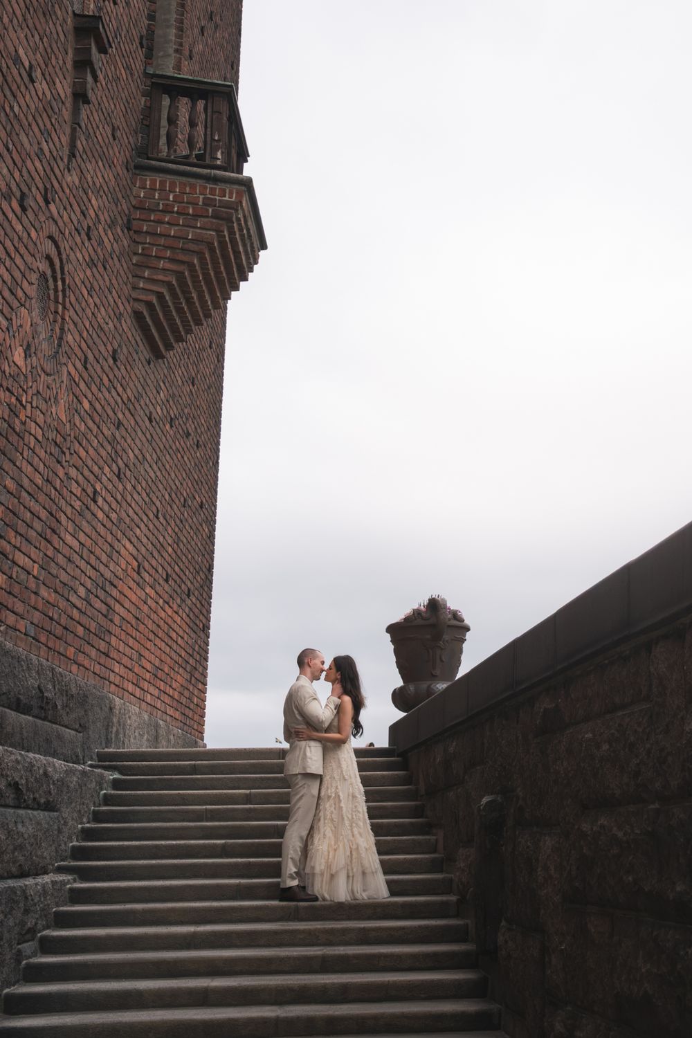 Bride and groom embrace on stone steps beside a historic brick castle under an overcast sky.