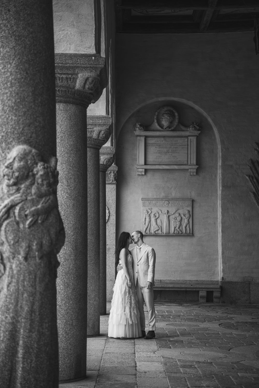 Black and white photo of a couple in wedding attire standing between classical columns in an elegant historic building.