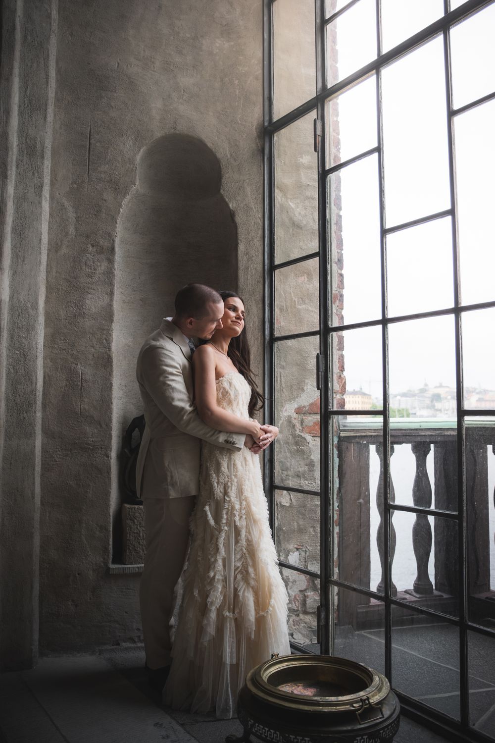 Bride and groom embrace by a large window in a rustic stone venue, bride wearing an elegant lace wedding dress.