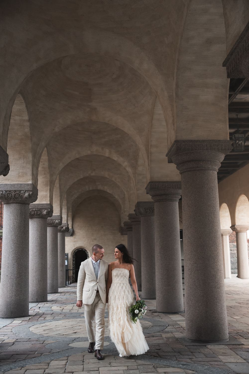 Bride and groom walk hand-in-hand through elegant stone archway colonnade on their wedding day.