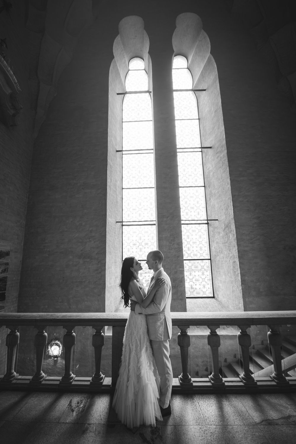 Black and white photo of a couple embracing before tall arched windows in an elegant building.