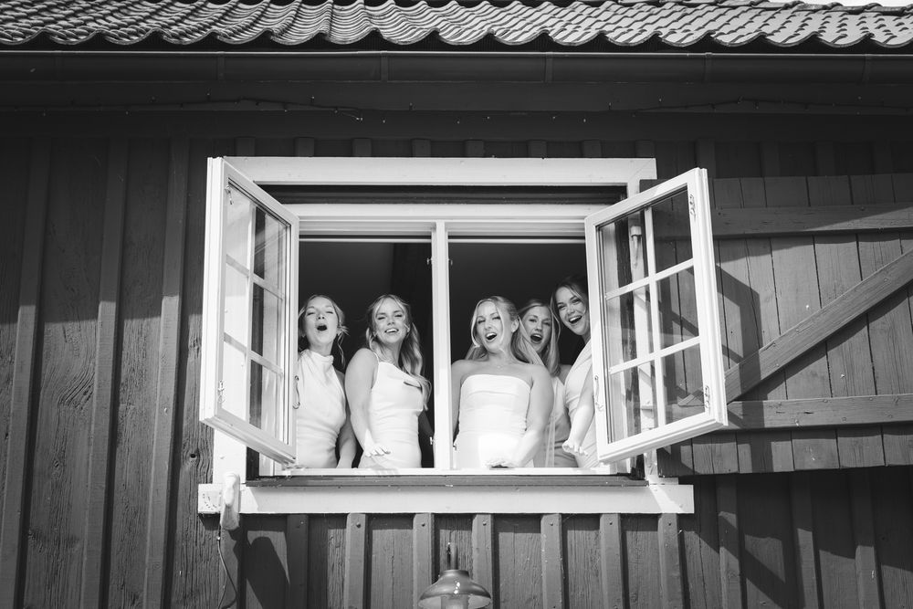 Black and white photo of a bride and bridesmaids laughing together in an open window of a rustic wooden building.
