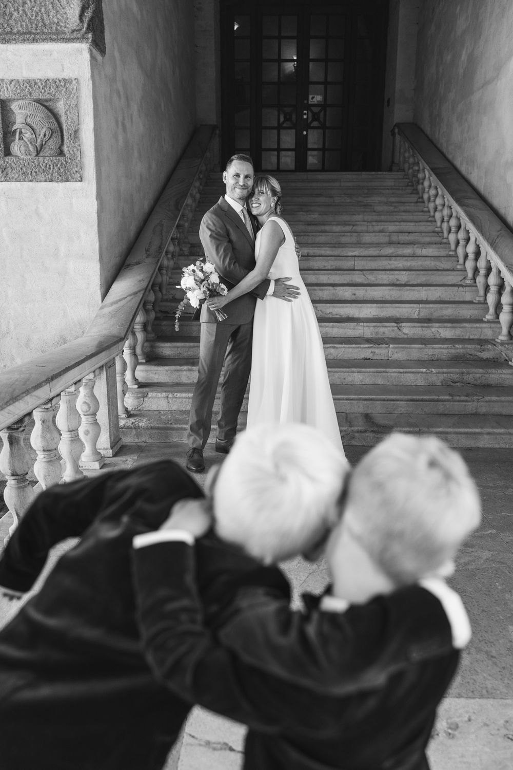 Black and white photo of a bride and groom embracing on grand staircase while photographer captures the moment.