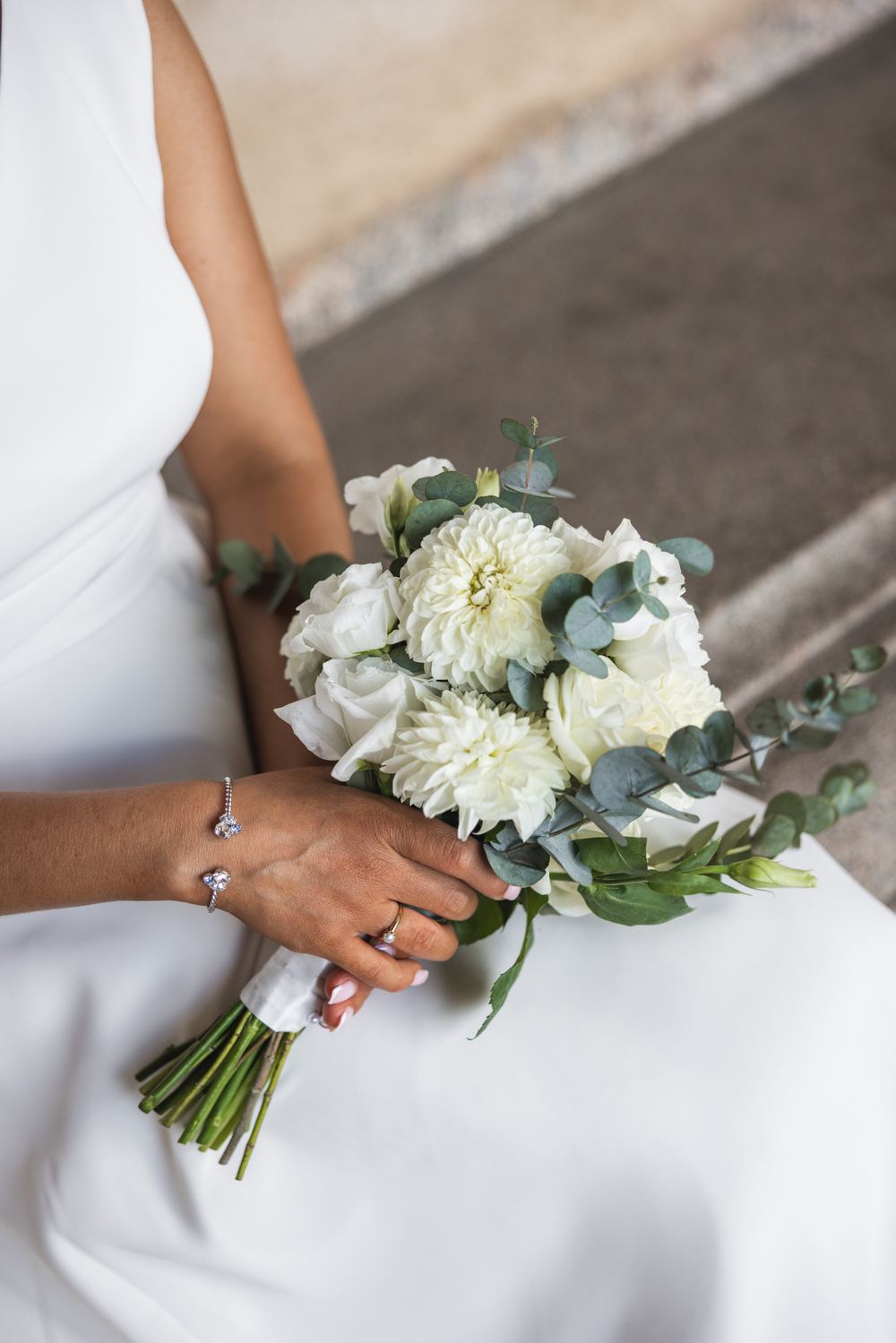 Bride in white dress holding elegant bridal bouquet of white dahlias, roses, and eucalyptus, wearing diamond bracelet and ring.
