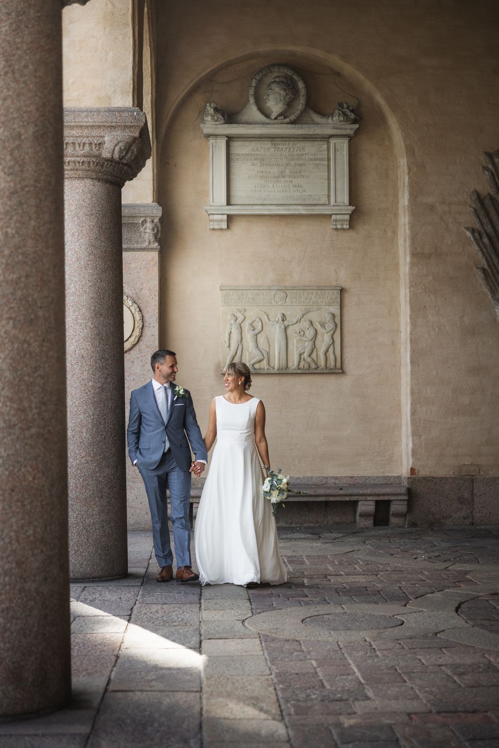 Bride and groom walk hand in hand through an elegant Italian courtyard with stone columns and classical relief sculptures.