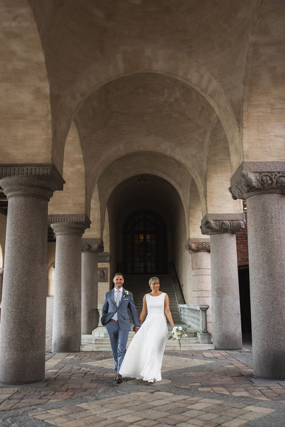 Bride and groom walking hand in hand through elegant stone archway with granite columns at their wedding venue.