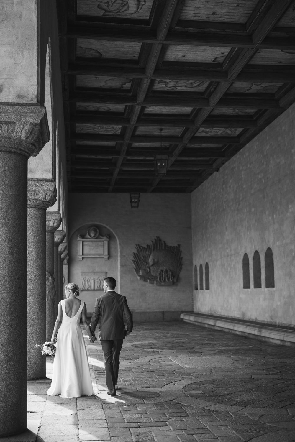 Black and white photo of bride and groom walking hand in hand through an elegant historic arcade with ornate ceiling.
