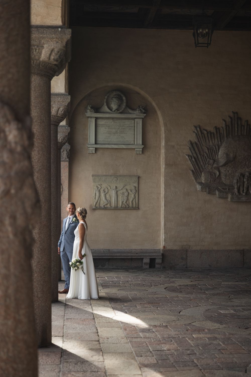 Bride and groom stand in elegant stone archway courtyard with classical relief sculptures during wedding ceremony.