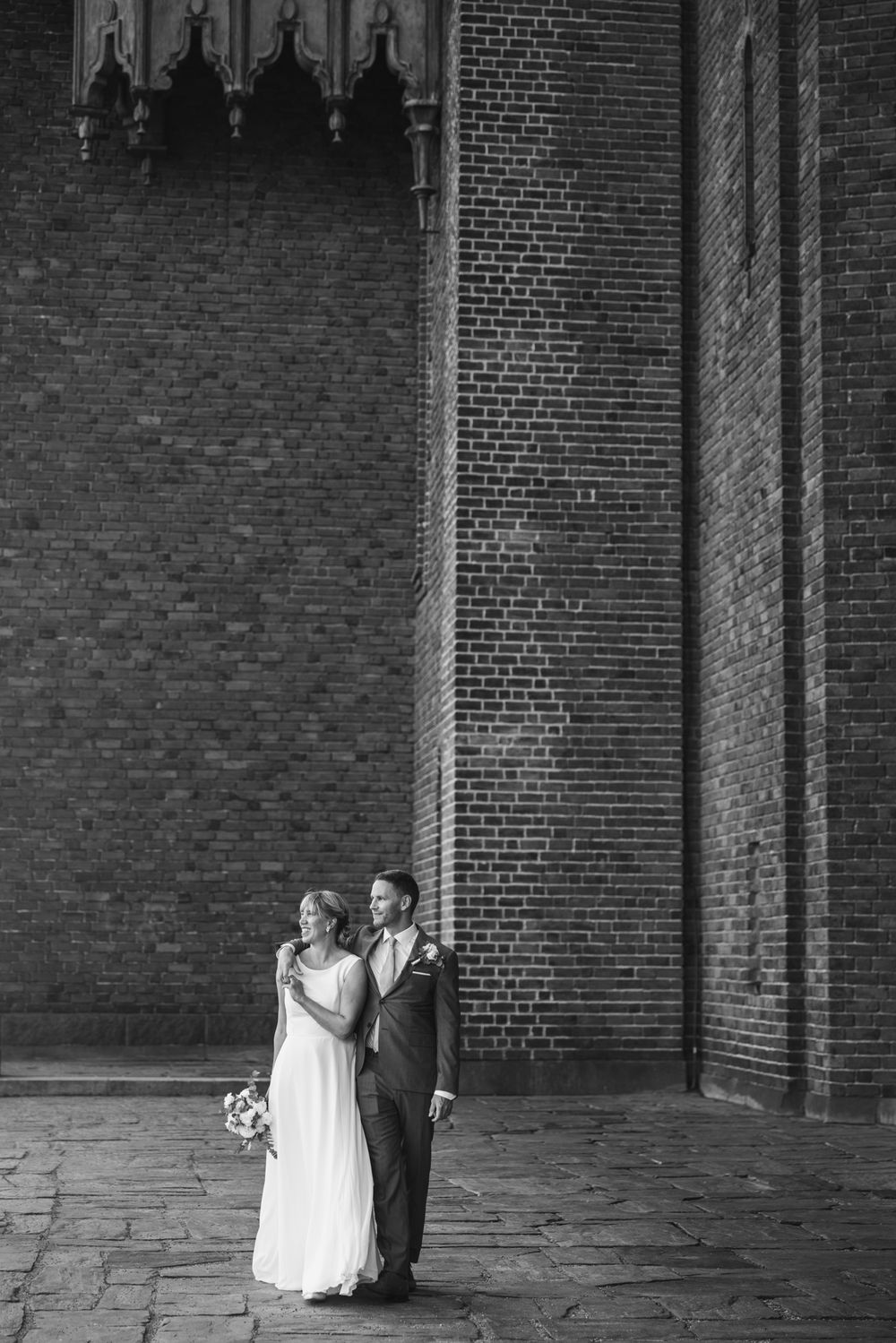 Black and white photo of a bride and groom standing together outside a historic brick church holding a bouquet.