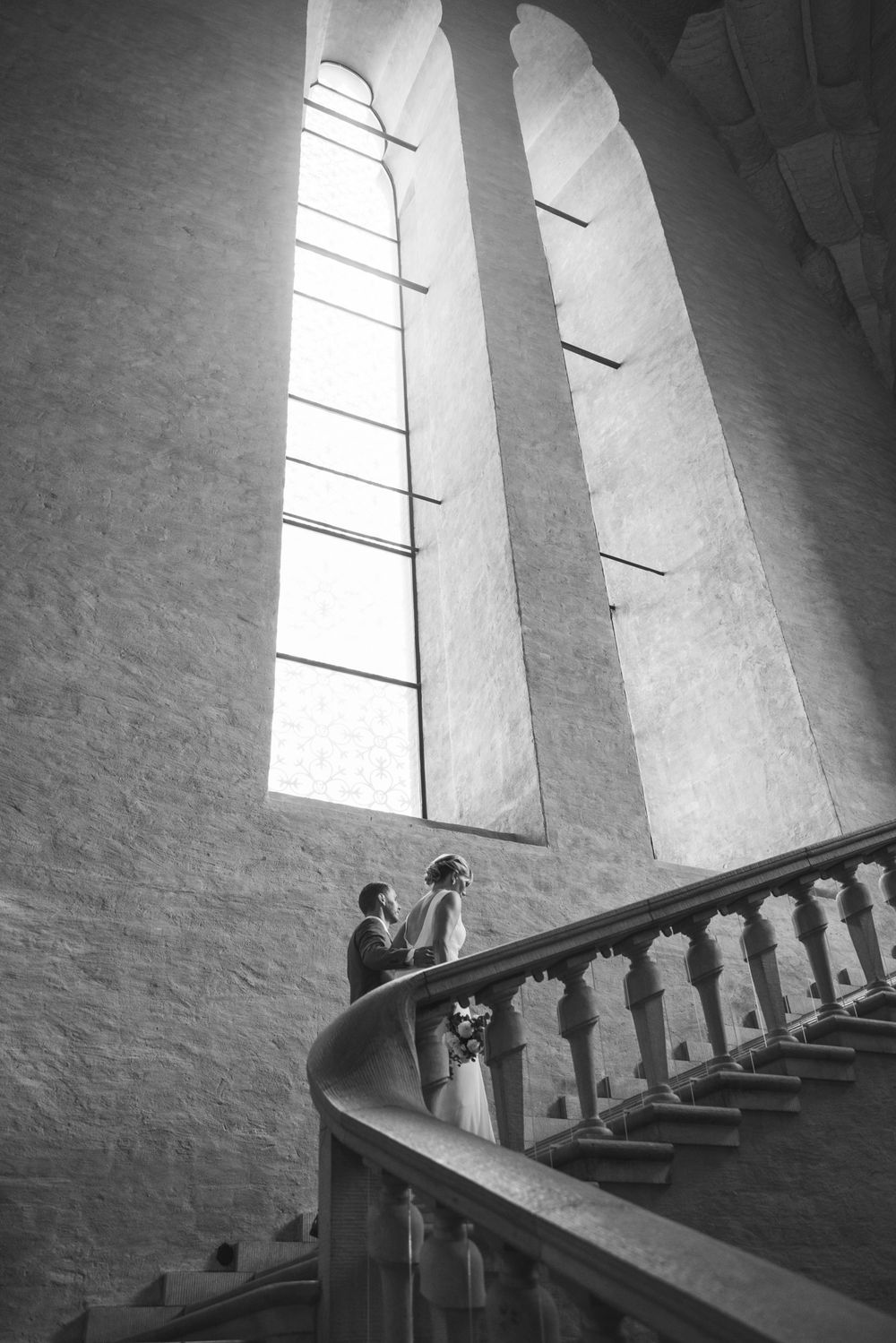Bride and groom share a romantic moment on a spiral staircase beneath tall arched church windows.