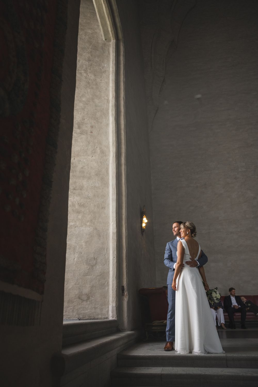 Bride and groom embrace on stone steps inside grand historic church with tall arched windows and warm lighting.