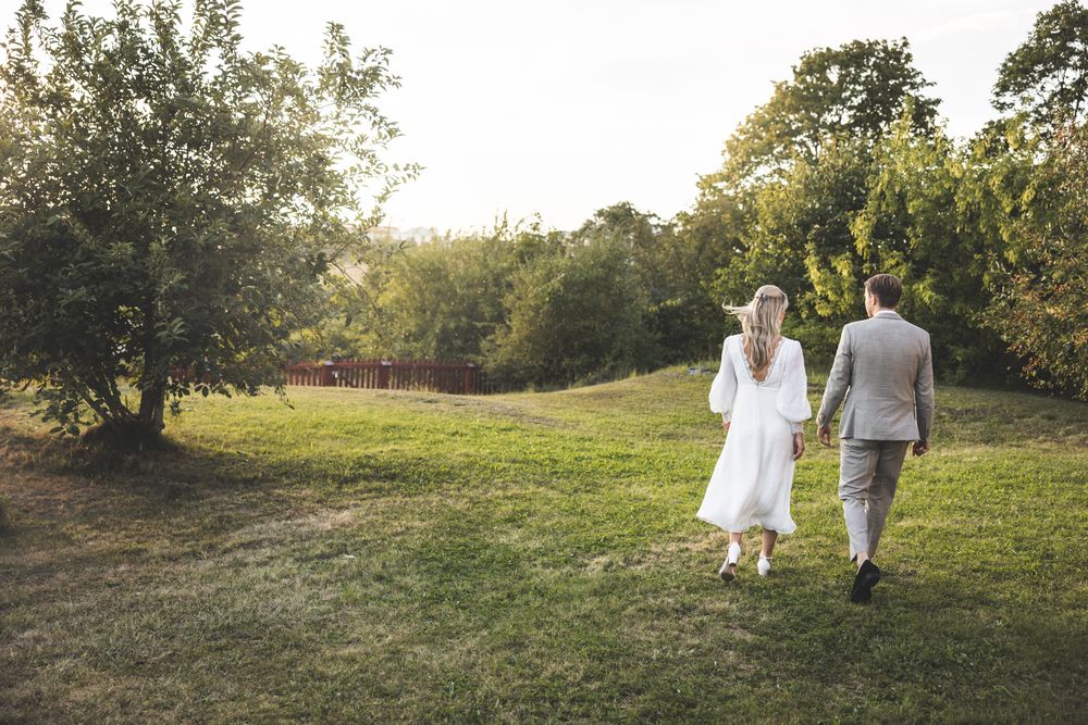 A couple in wedding attire walks through a sunlit meadow holding hands during a romantic sunset photoshoot.