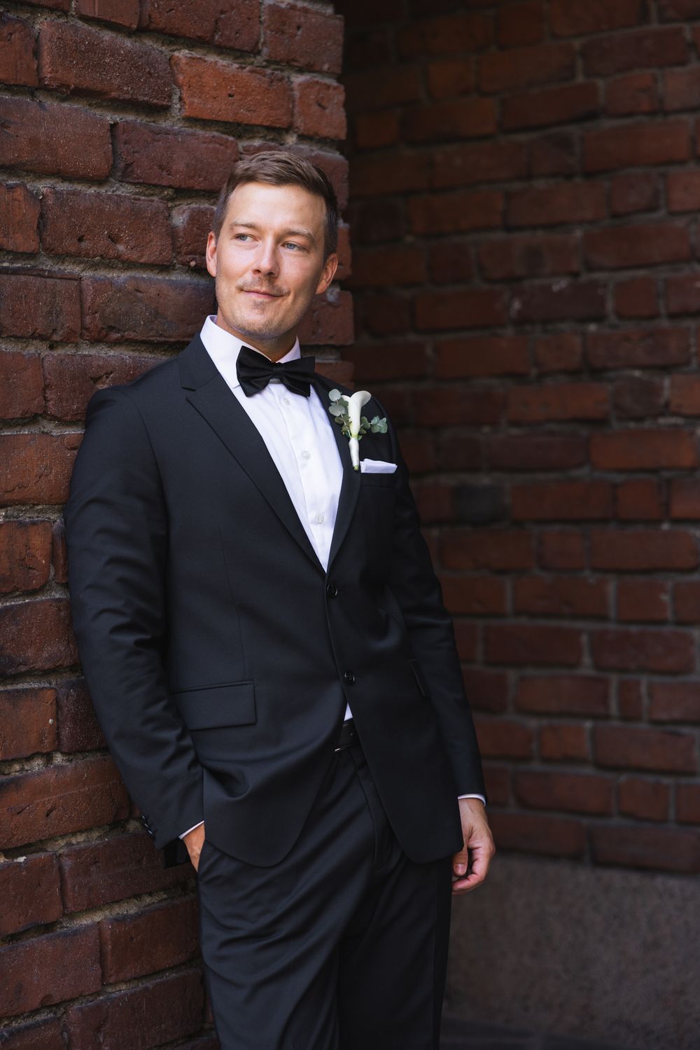 Groom in black tuxedo with bow tie and boutonniere leaning against red brick wall.