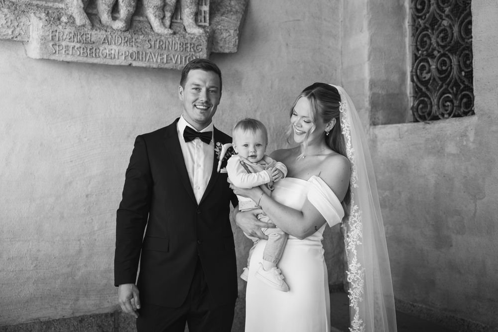 Black and white wedding photo of a couple in formal attire holding their baby, posing in front of a stone wall.