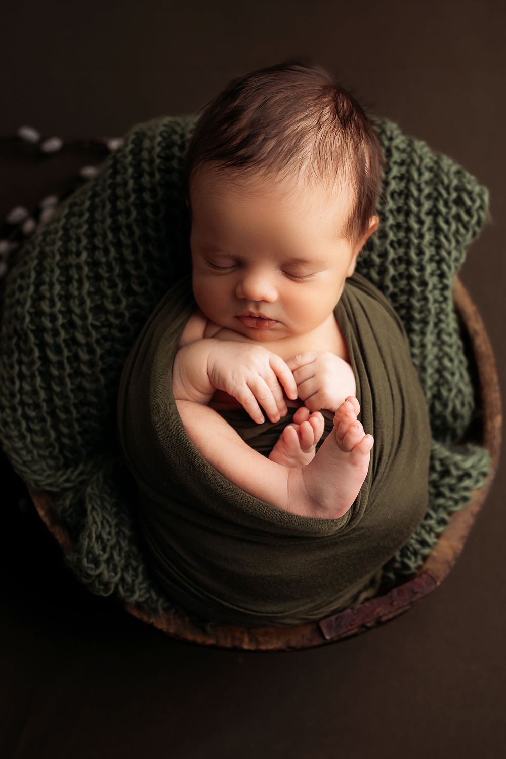 Newborn sleeping peacefully on dark backdrop wrapped in green knit blanket with hands near face.