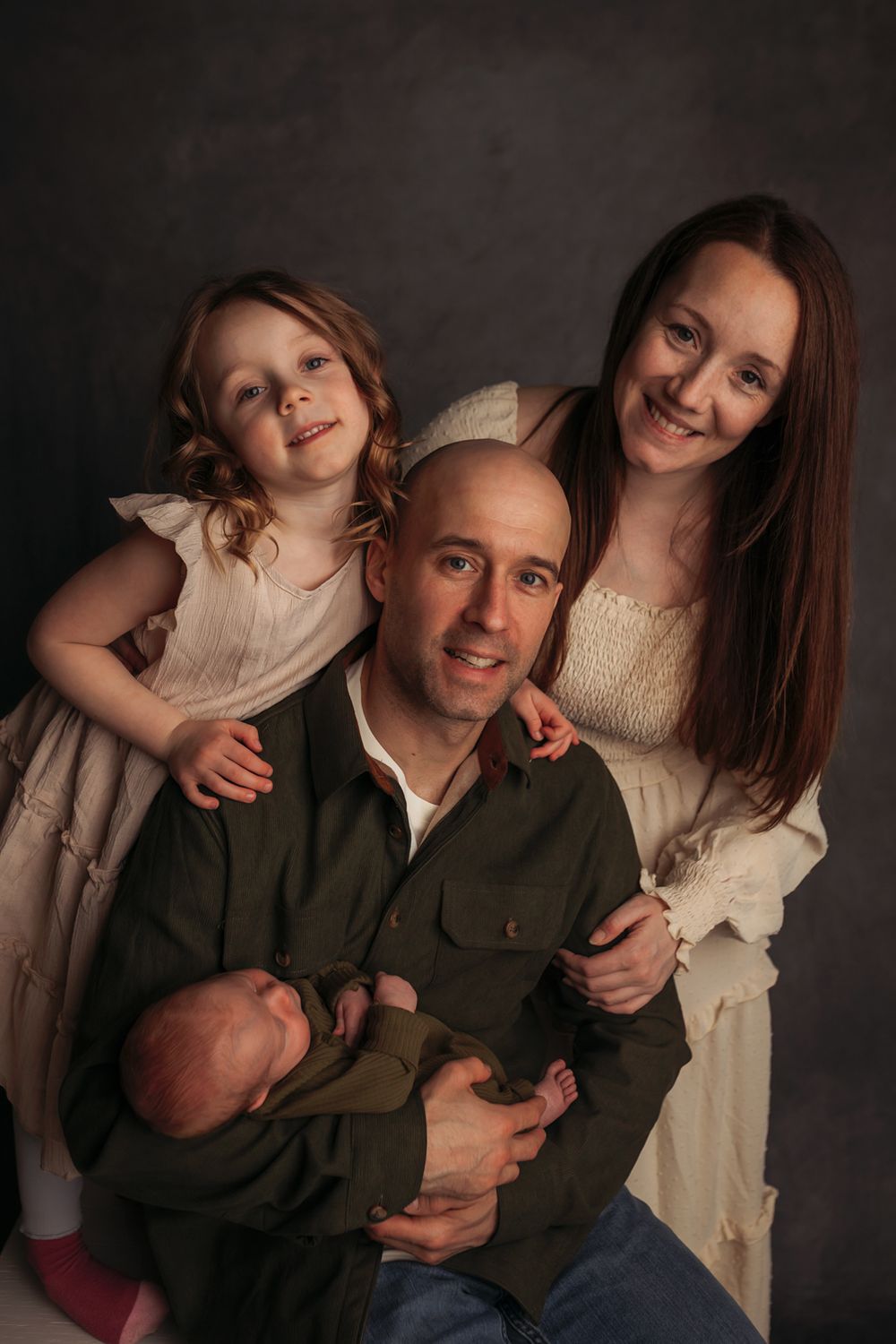 Family portrait of parents with young daughter and newborn baby against a dark studio background.