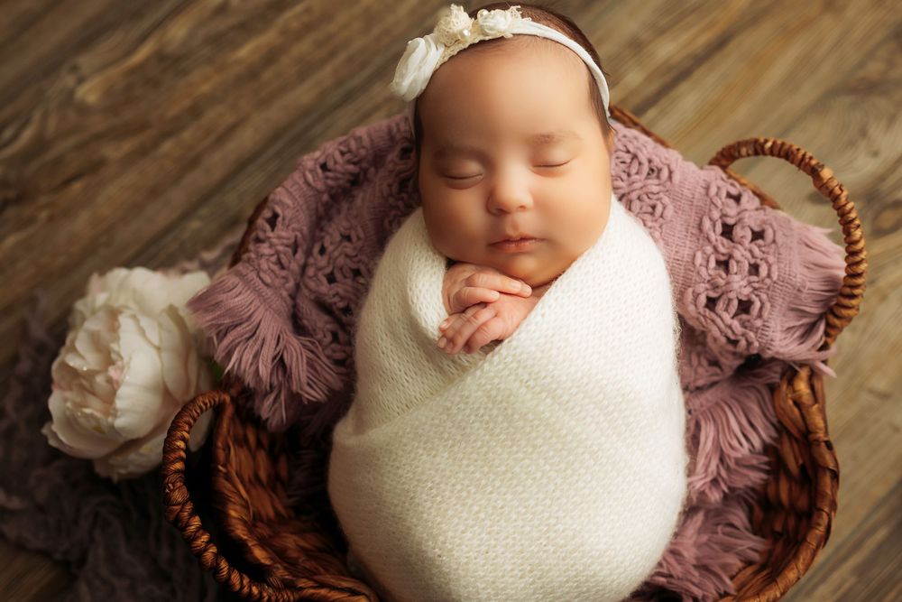 Newborn baby wrapped in white blanket lying on brown wicker basket with purple fabric background.