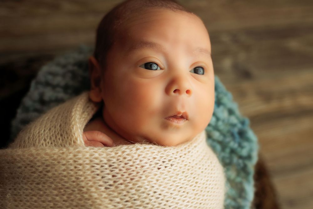 Close-up portrait of newborn baby swaddled in cream-colored wrap against wooden backdrop.