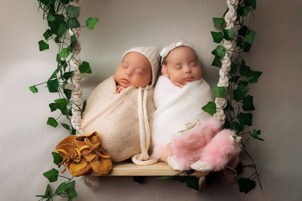 Newborn twins in cream wraps sleeping peacefully on wooden shelf with green vine decorations.