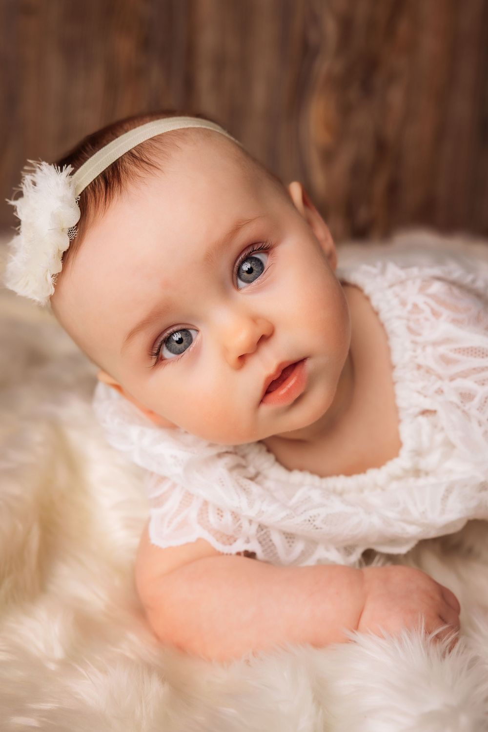 Adorable baby girl with blue eyes wearing a white lace outfit and floral headband lying on a fluffy white fur rug.