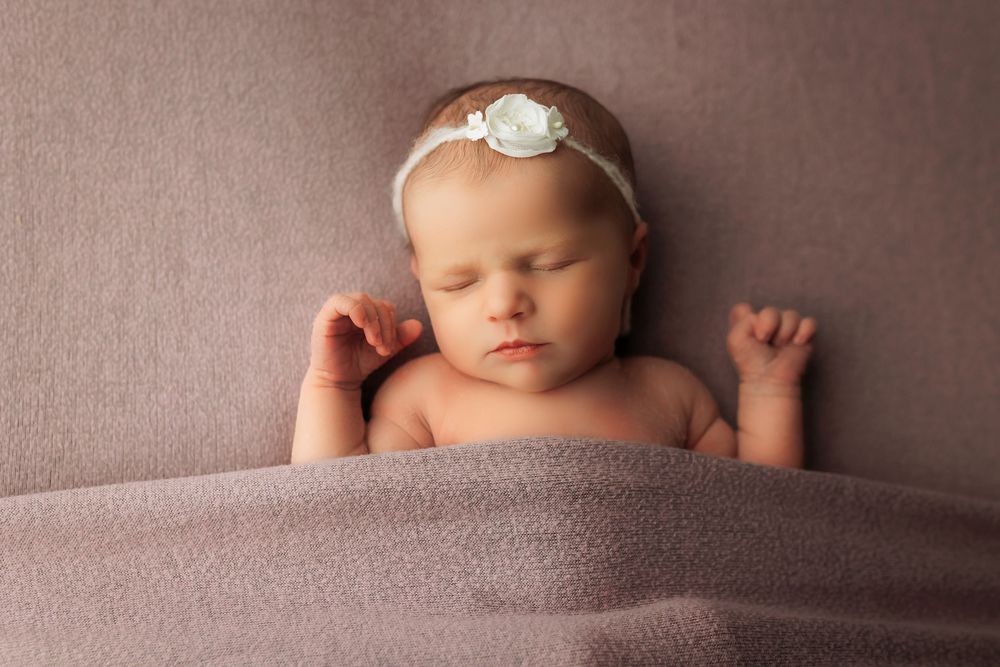 Sweet newborn wearing white headband nestled in cozy mauve blanket during studio photography session.