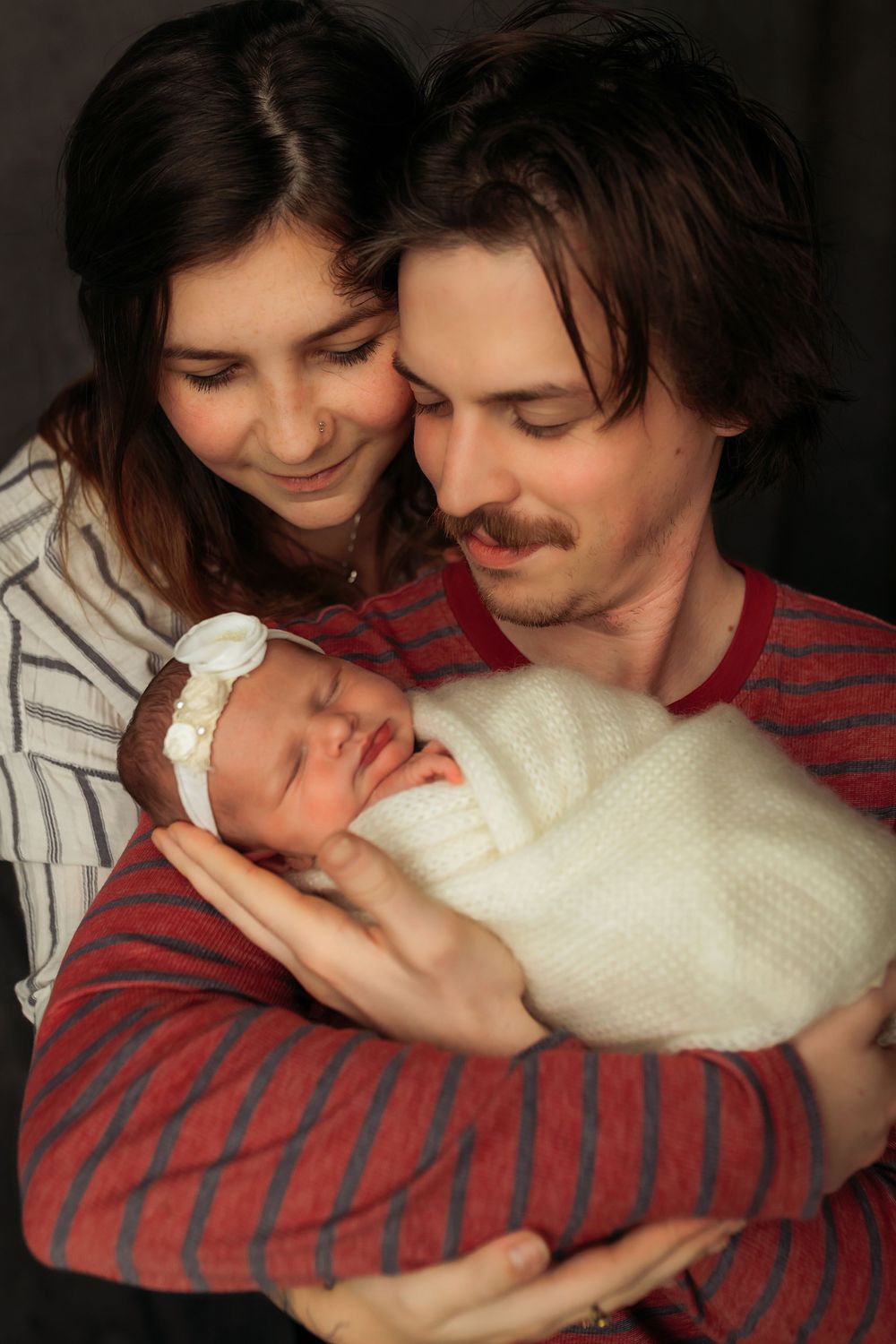 Parents sharing intimate moment with newborn baby against dark studio backdrop.