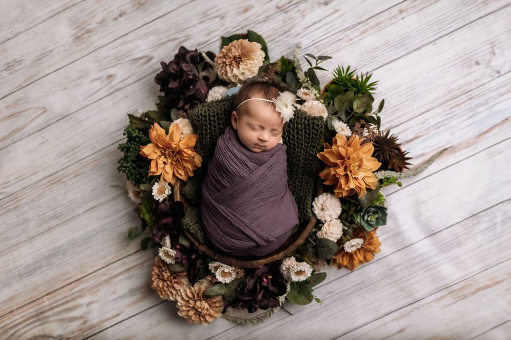 Sleeping newborn wrapped in purple swaddle surrounded by autumn floral wreath on wooden floor.