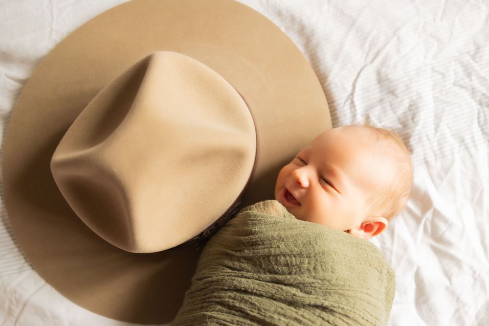 Sleeping newborn swaddled in green wrap next to a tan cowboy hat.