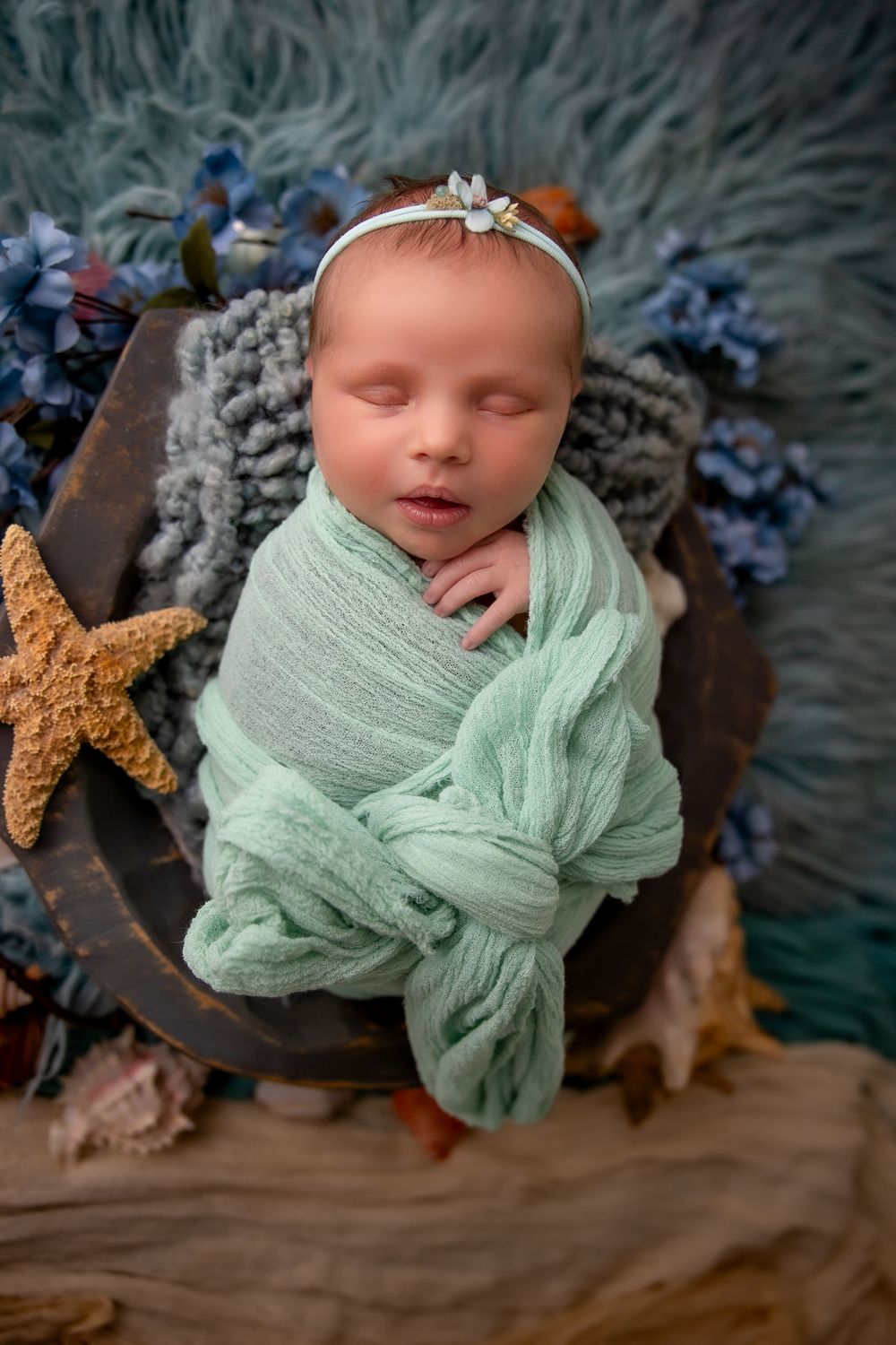 Newborn wrapped in mint green swaddle sleeps peacefully in wooden bowl with starfish and blue flower decorations.