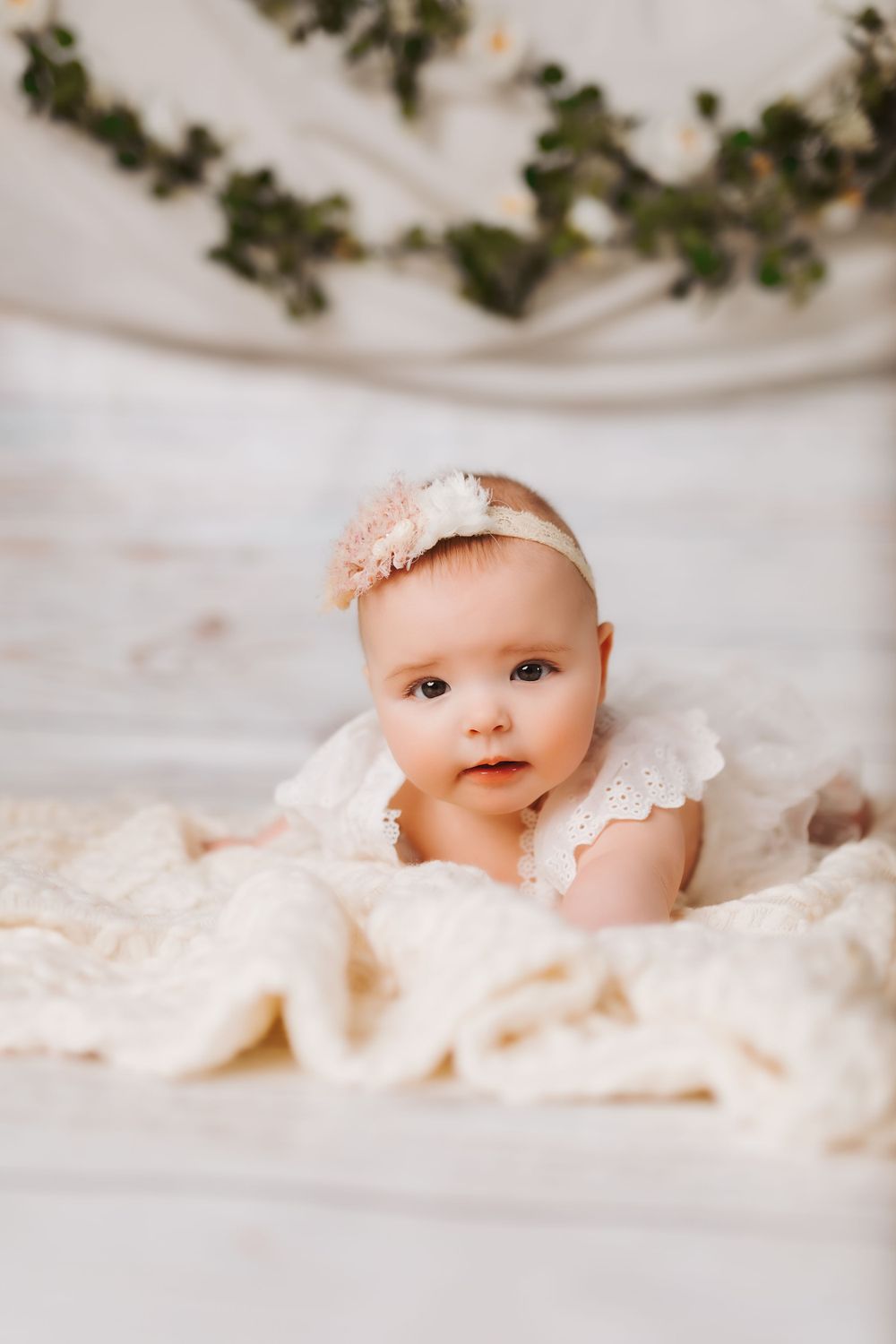 Baby in delicate white lace dress lying on cream fabric with green ivy backdrop.
