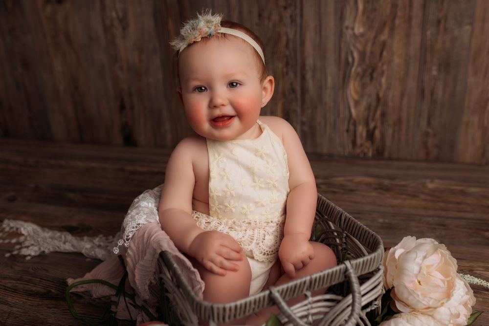 Smiling baby girl in cream lace romper sitting in wicker basket on rustic wood background with peony flower.
