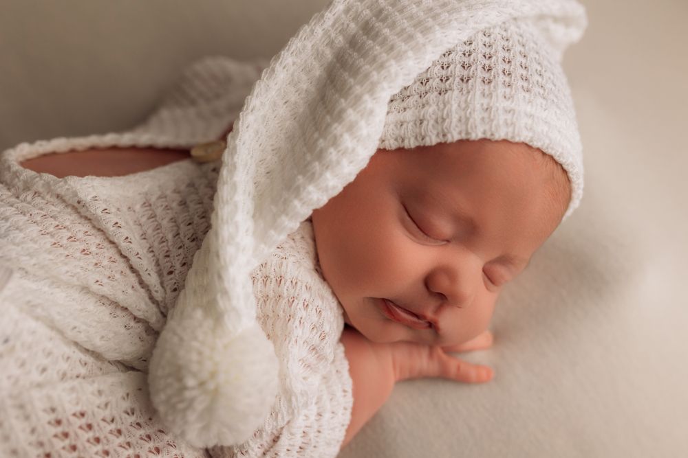 Sleeping newborn baby in white knit hat and outfit posed on soft white blanket.