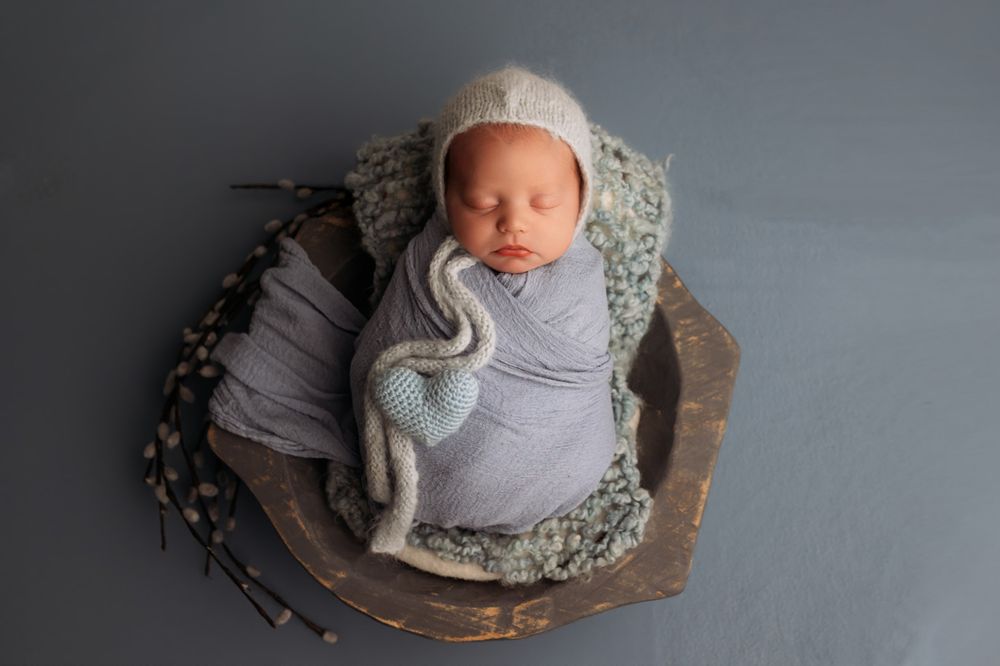 Swaddled newborn baby in gray wrap resting in wooden bowl with floral accents.