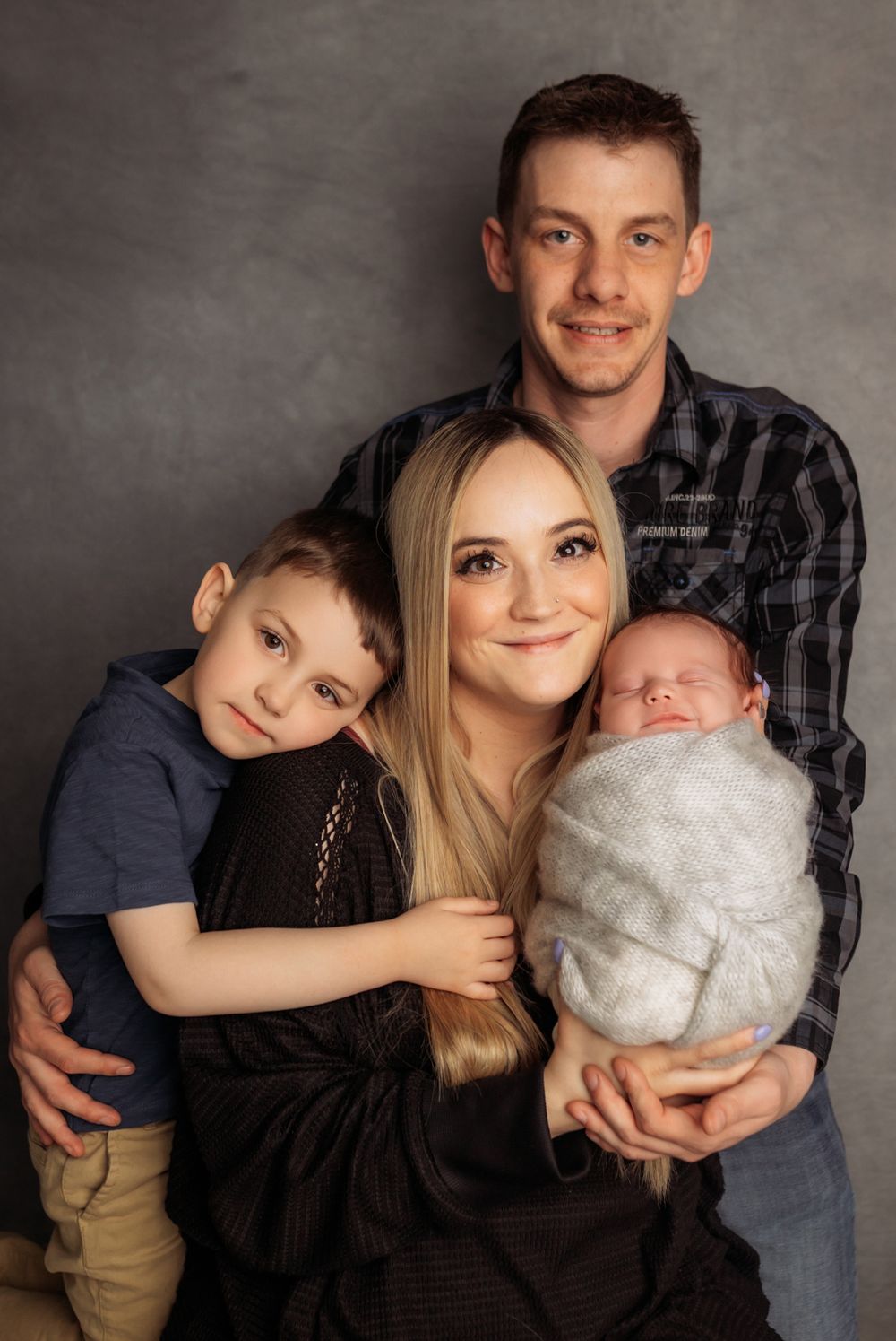 Family portrait with parents, toddler son, and swaddled newborn baby on gray background.