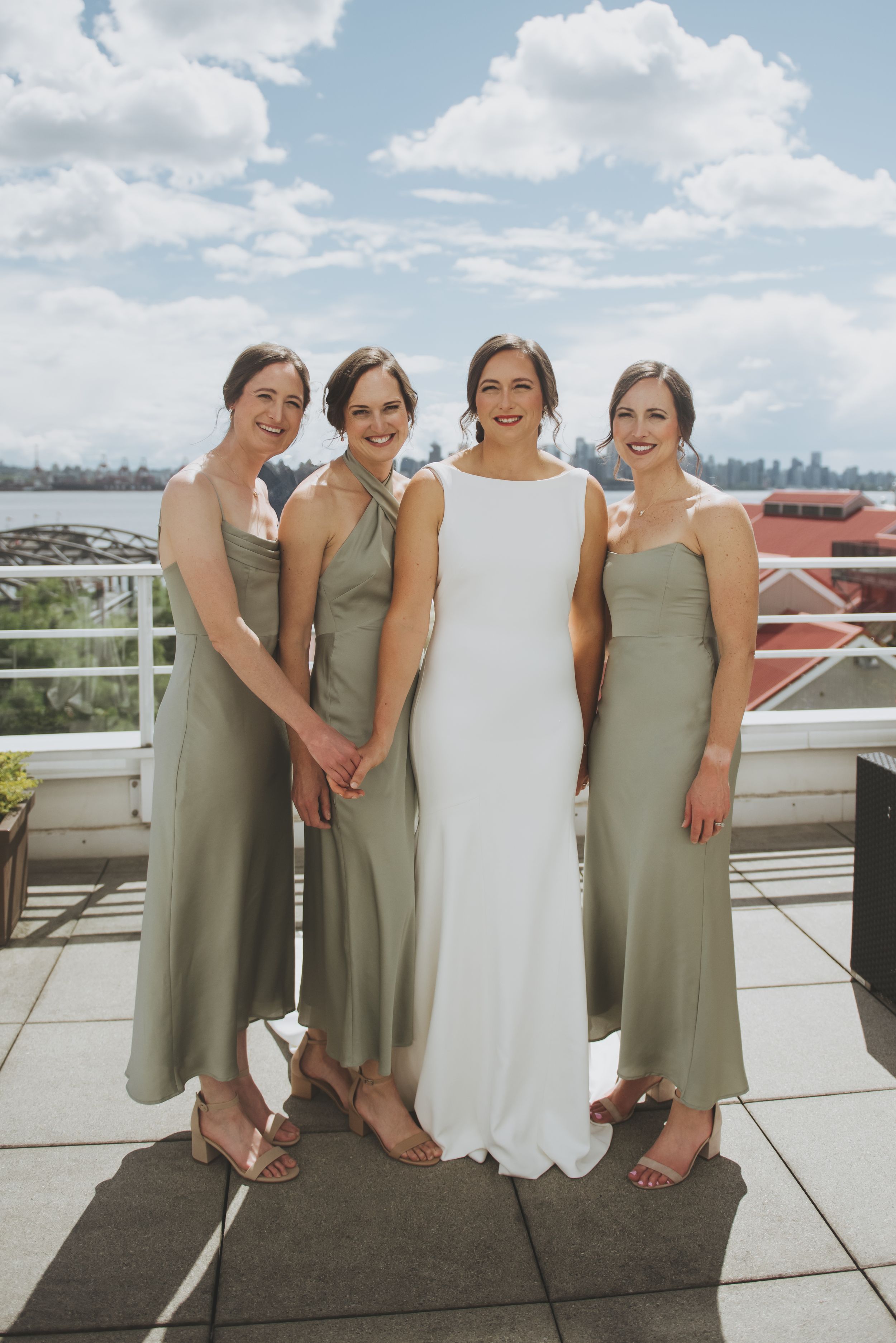 Bridesmaids in sage green dresses pose with bride in white on a rooftop terrace.