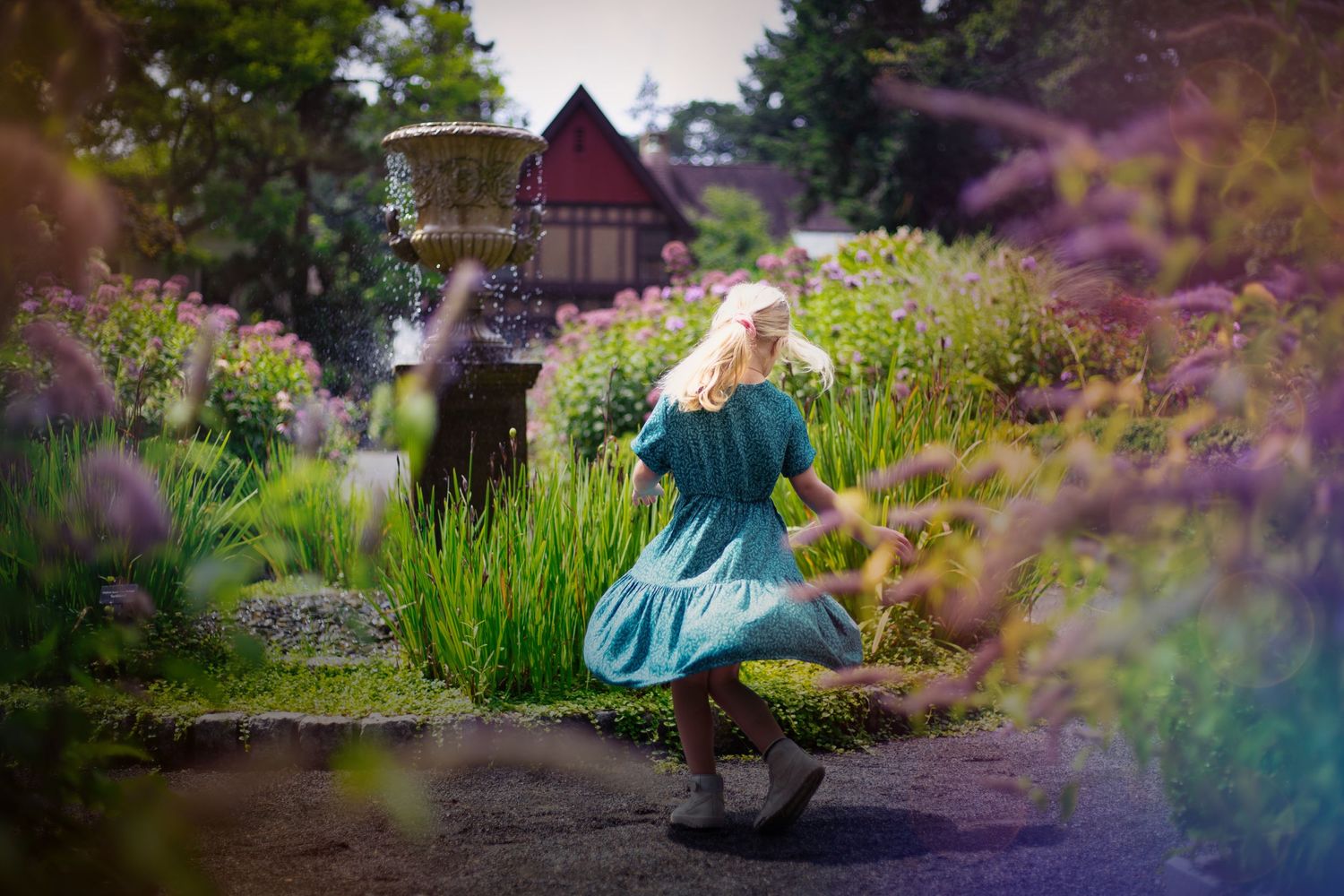 Figure in teal dress twirls in magical garden setting with flowers and red barn in background.