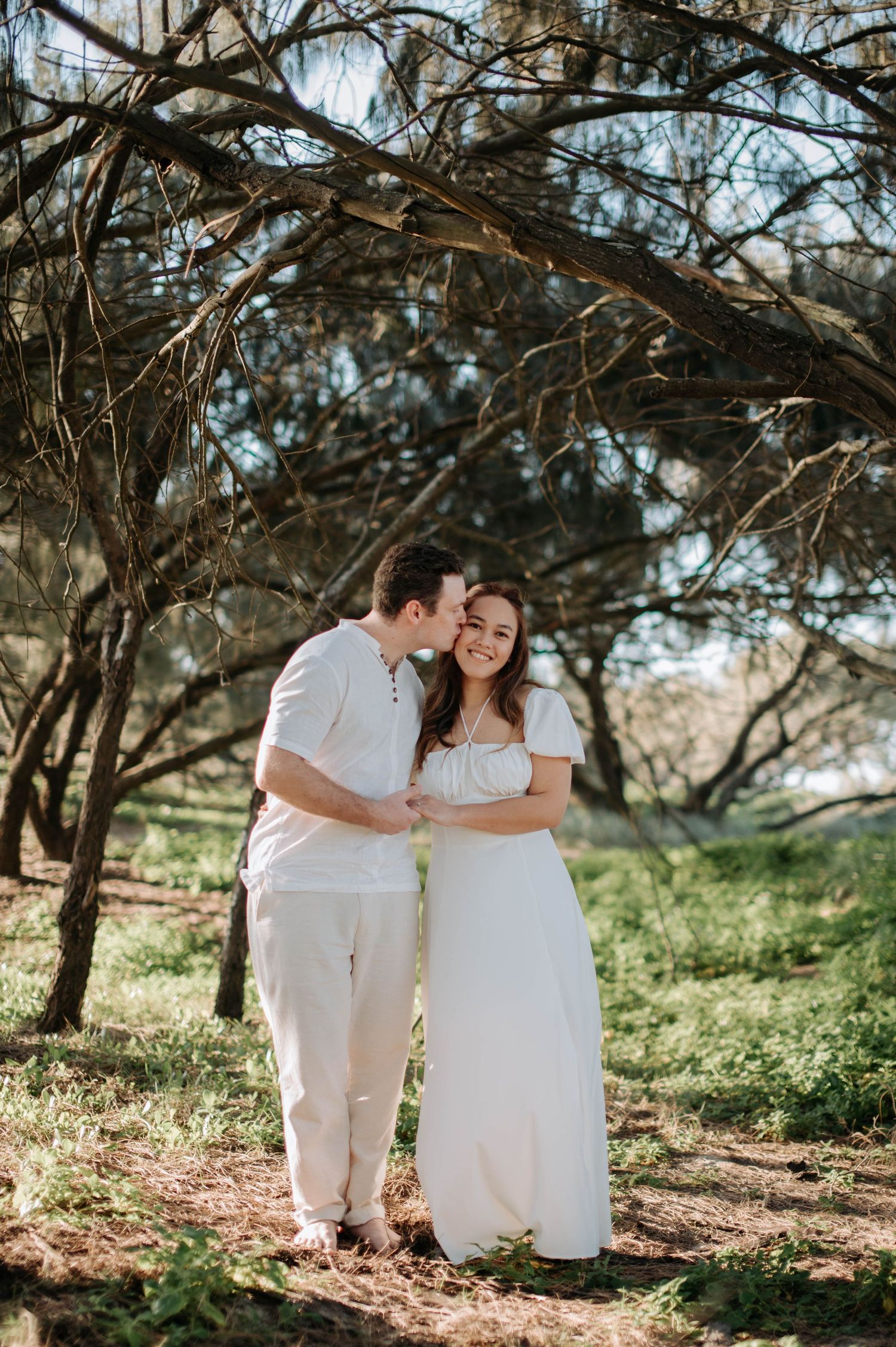 Couple in all white attire embracing under tree branches in a natural outdoor setting.
