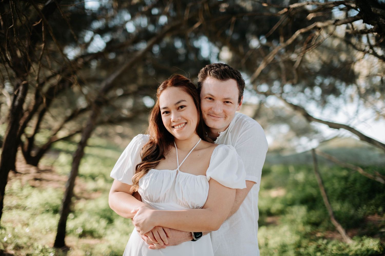 Romantic engagement photo of couple in white clothing sharing an intimate moment outdoors.